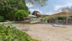 An outdoor seating area with tables and chairs, surrounded by trees and a gravel path. - Les crouquets - Le Pêcher (Jayac)