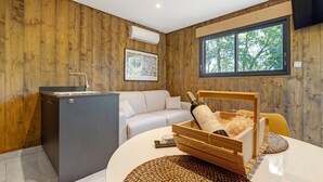 A room with wooden walls, a white sofa, a small kitchen area, and a large window with a view of trees.
