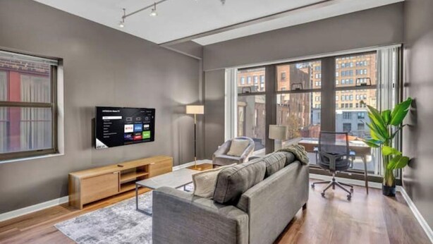 A modern living room with a grey sofa, a TV mounted on the wall, a wooden entertainment unit, and a large window offering a city view. - Playhouse Square Condo /  Free Parking / Downtown (Cleveland)