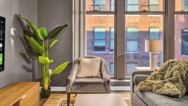 Een moderne woonkamer met een grijze fauteuil, een grijze bank, een houten bijzettafeltje en een grote groene plant.