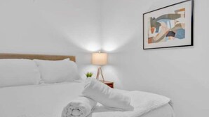 Una cama bien tendida con sábanas blancas, una lámpara en la mesita de noche y un cuadro abstracto enmarcado en la pared.