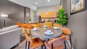 A modern kitchen with wooden cabinets, a dining area with a round table and orange chairs, and a large potted plant.