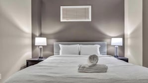 A neatly made bed with a rolled towel, two bedside tables with lamps, and a window with blinds.