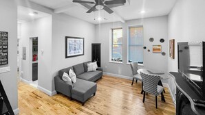 A modern living room with a gray sectional sofa, a ceiling fan, a dining area with a small table and chairs, and large windows with blinds. - Exquisite 1st Floor One Bedroom in Historic Marble Hill w/ Modern Amenities (Baltimore)