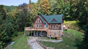 A large wooden house with a green roof, a wooden deck, and a staircase leading to the main entrance. - Spacious 4-bedroom + loft, 8 Min Drive to Resort, Equipped Hot Tub & Activities (Cass)