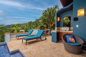 Zona junto a la piscina con sillones de descanso, una estación de barbacoa y vistas de la vegetación circundante.