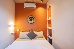 A hotel room with a bed, an air conditioner, a wall-mounted decorative plate, and a shelf with items.