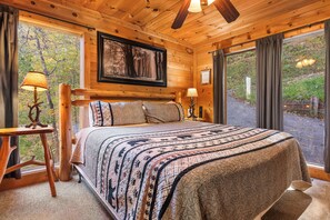 Un dormitorio de cabaña de madera con una cama grande, dos lámparas de noche, un cuadro enmarcado en la pared y una ventana con cortinas.