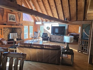 L’intérieur chaleureux d’un chalet avec une cheminée en pierre, des poutres en bois, un grand téléviseur, des meubles en cuir et un escalier.