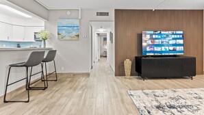 Eine moderne Küche mit weißen Schränken und einer grauen Arbeitsplatte, ein Essbereich mit zwei grauen Stühlen, ein Flachbildfernseher auf einem schwarzen Schrank und eine Wand mit Holzlatten.