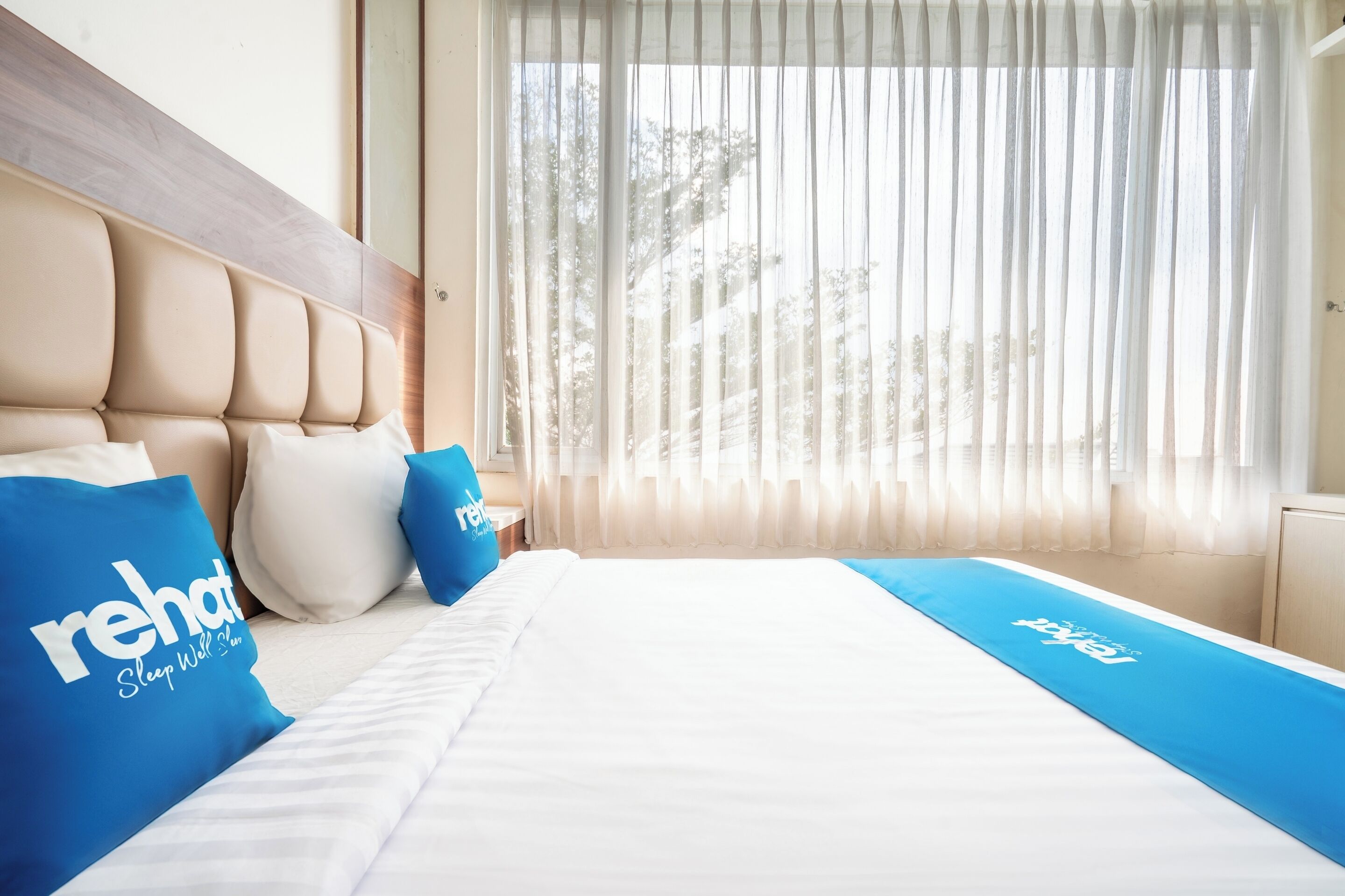 Une chambre d’hôtel avec un lit, une tête de lit, une fenêtre avec des rideaux légers et un couvre-lit bleu orné d’un motif.