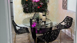 Un coin repas avec une table en verre entourée de chaises noires aux pieds dorés, un néon mural et une composition florale décorative.