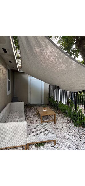 A covered outdoor seating area with wicker furniture and a wooden table, surrounded by white pebbles and a fence. - Live the Miami life in style and comfort (Miami)