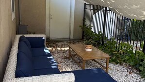 Un canapé en osier et une petite table placés sous un espace ombragé, avec une porte et une clôture en métal en arrière-plan.