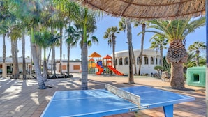 An outdoor area with a ping pong table, palm trees, a playground with a slide, and a building with arches. - Vacation Apartment 'Brisa Azul' with Shared Pool, Private Terrace and Wi-Fi (Corralejo)