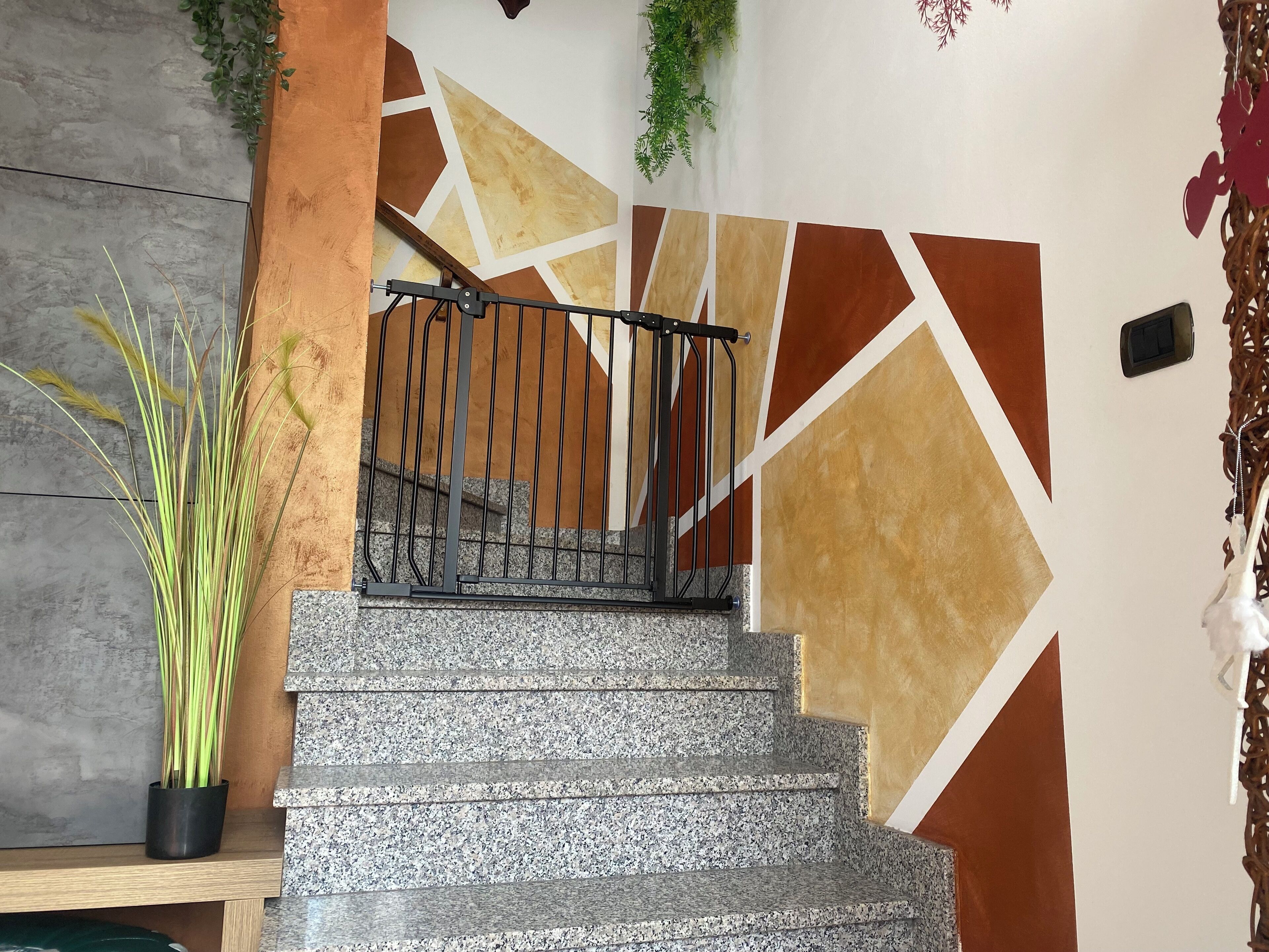 Una escalera con un diseño geométrico en la pared, barandilla de metal y una planta en maceta.