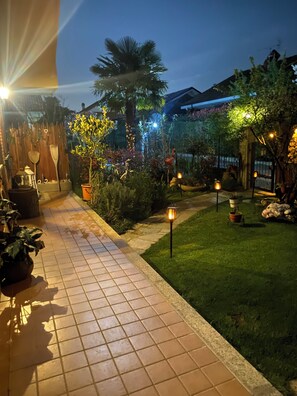 A well-lit garden pathway with potted plants, a flamingo statue, and decorative lights.