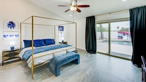A modern bedroom with a four-poster bed, a bench, a ceiling fan, and a view of a swimming pool.