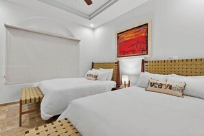 Une chambre d’hôtel avec deux lits, une fenêtre avec un store, une tête de lit en bois, un tableau mural et un banc en osier.