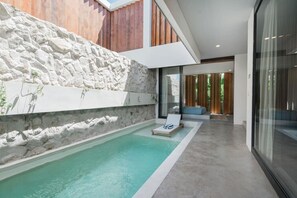 A modern outdoor pool area with a stone wall, a lounge chair, and a glass door leading to a wooden deck. - Stylish 2BR Villa Private Pool (Kuta Utara)