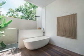 A modern bathroom with a freestanding bathtub, a wooden floor, and a view of greenery outside. - Stylish 2BR Villa Private Pool (Kuta Utara)