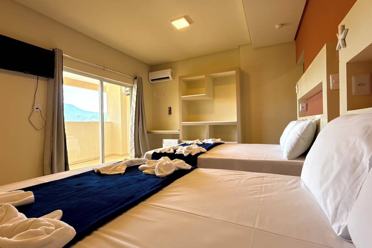 Quarto de hotel com duas camas, uma televisão fixada na parede e uma porta de vidro deslizante para uma varanda.