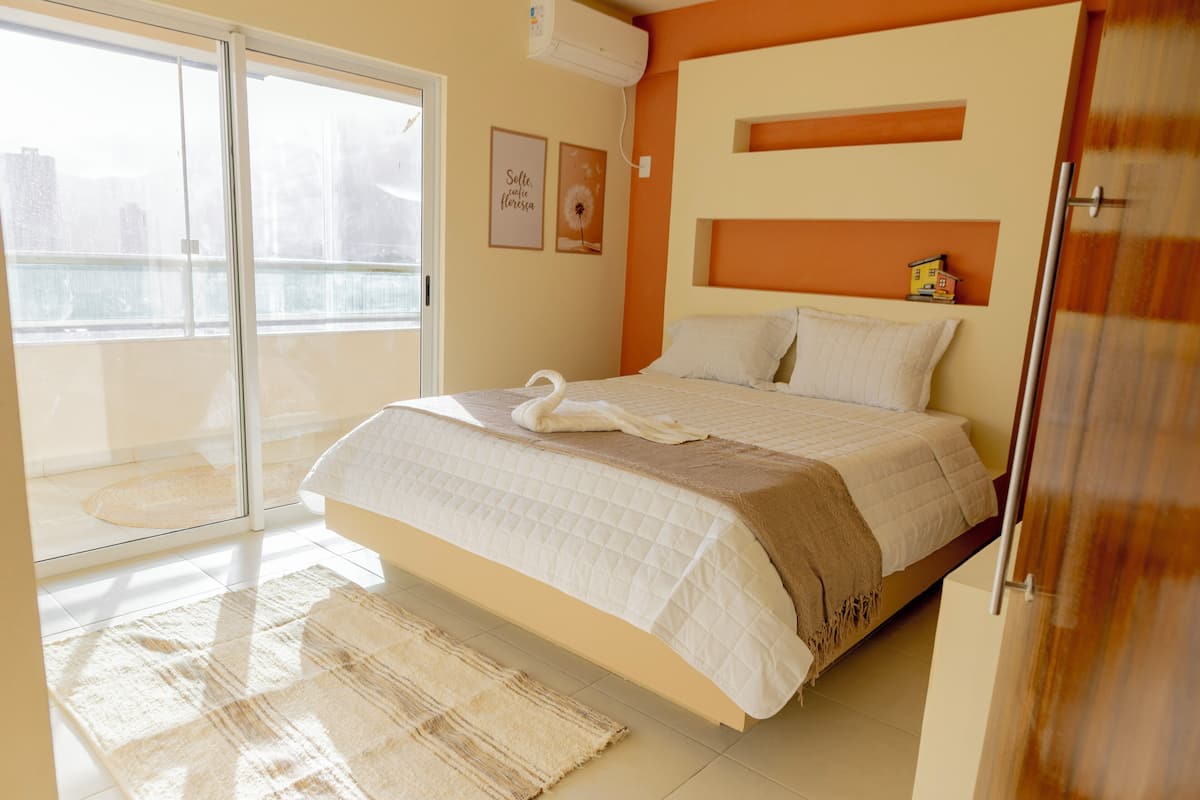 Quarto com cama, porta de vidro deslizante para uma varanda e uma pequena prateleira com uma casinha de brinquedo.