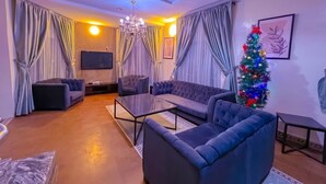 Una sala de estar moderna con un sofá modular, una mesa de centro de vidrio y un televisor montado en la pared. Se ve un árbol de Navidad en una esquina, y la habitación tiene un ambiente cálido gracias a las cortinas y una araña.