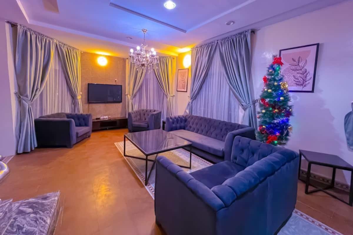 A modern living room with a sectional sofa, a glass coffee table, and a TV mounted on the wall. A Christmas tree is visible in the corner, and the room has a warm ambiance with curtains and a chandelier.