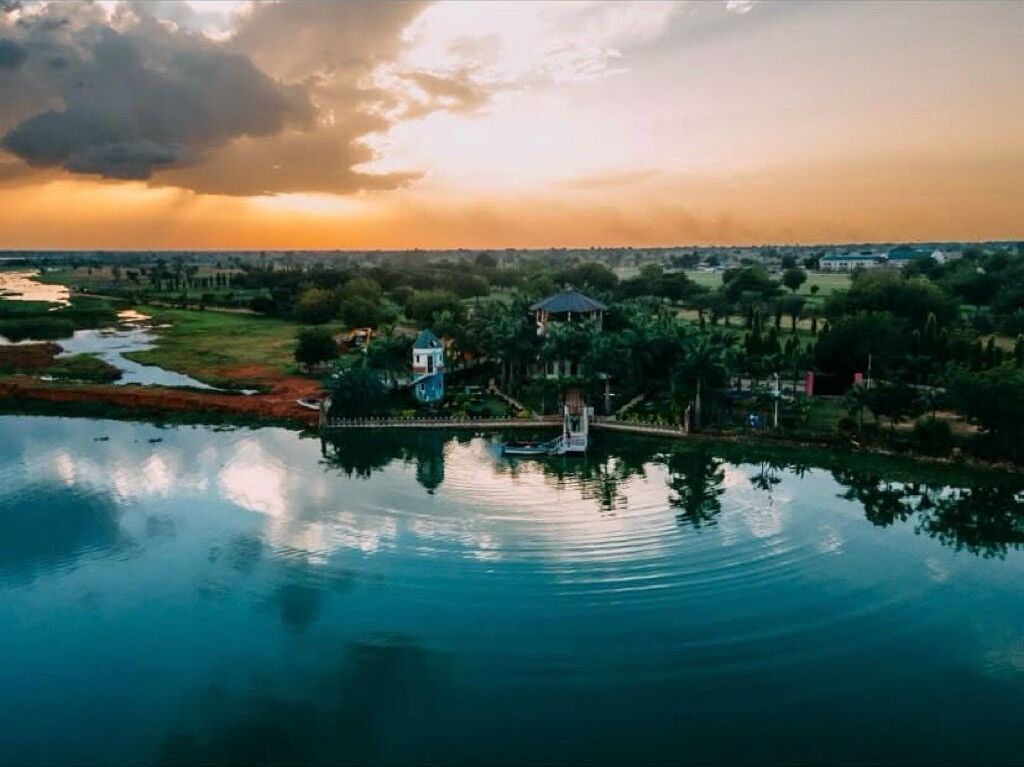 A serene lakeside view with a gazebo and a small boat, surrounded by lush greenery and a distant cityscape.