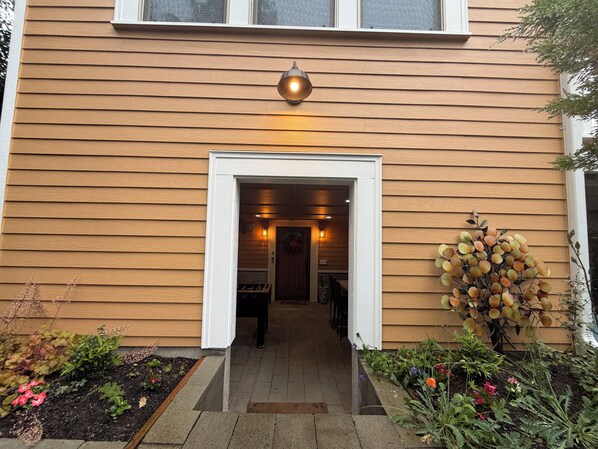 Una casa de madera con un marco blanco en la entrada, una lámpara sobre la puerta y un jardín con diversas plantas y flores.
