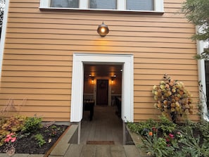 A wooden house with a white-framed entrance, a light fixture above the door, and a garden with various plants and flowers.