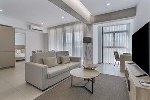 Ein modernes Wohnzimmer mit einer Couch, zwei Beistelltischen, einer Stehlampe, einem Fernseher und einem Essbereich mit Stuhl und Tisch.