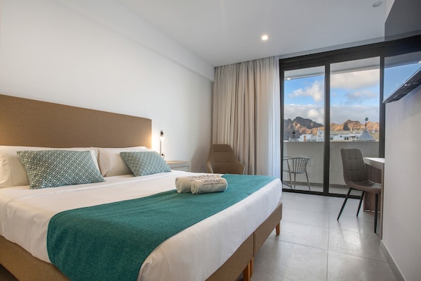 A hotel room with a bed, a chair, a balcony, and a view of the city.