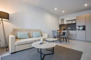 Un salon moderne comprenant un canapé, deux tables d’appoint, une lampe, ainsi qu’un coin cuisine avec une table et des chaises.