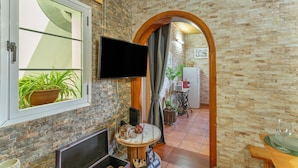 Une pièce avec un mur en briques, un téléviseur à écran plat fixé au mur, une table ronde ornée d’une plante et une porte en arc donnant sur une autre pièce.