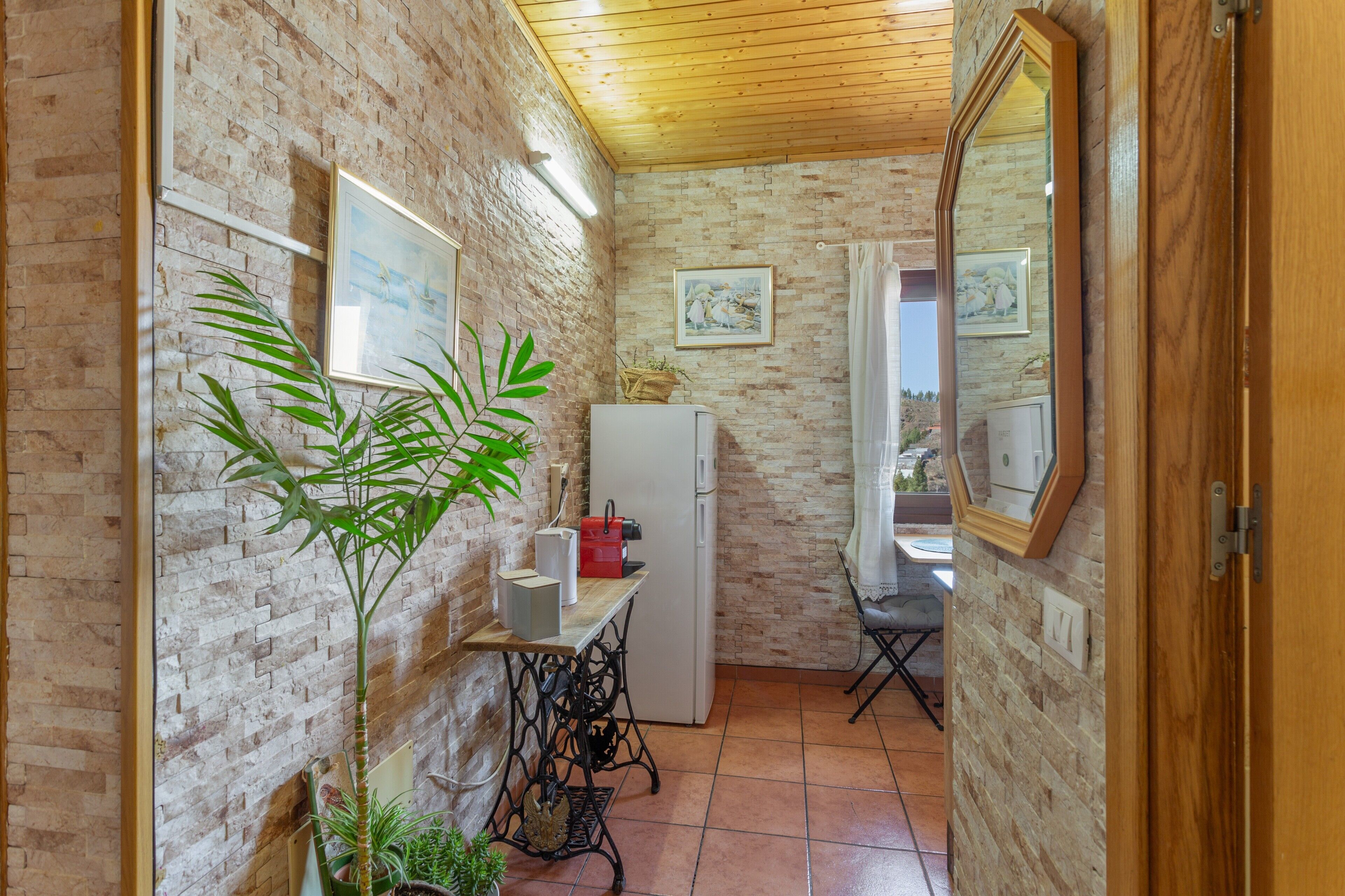 Cozinha compacta com parede de tijolos, geladeira, mesa com uma planta e um espelho.