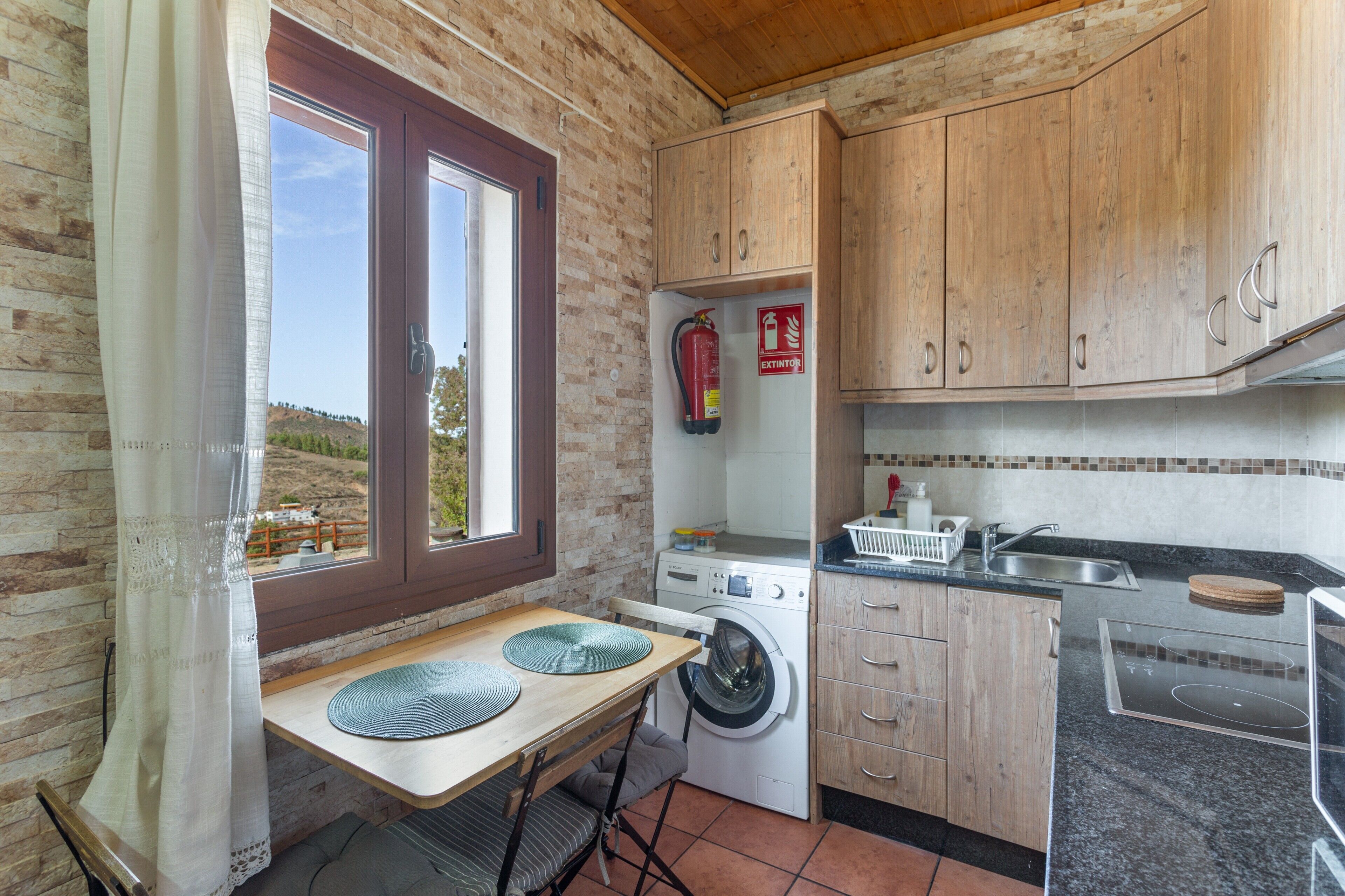 Eine kompakte Küche mit Waschmaschine, Holzschränken, einem Spülbecken und einem Fenster mit Blick auf eine hügelige Landschaft.