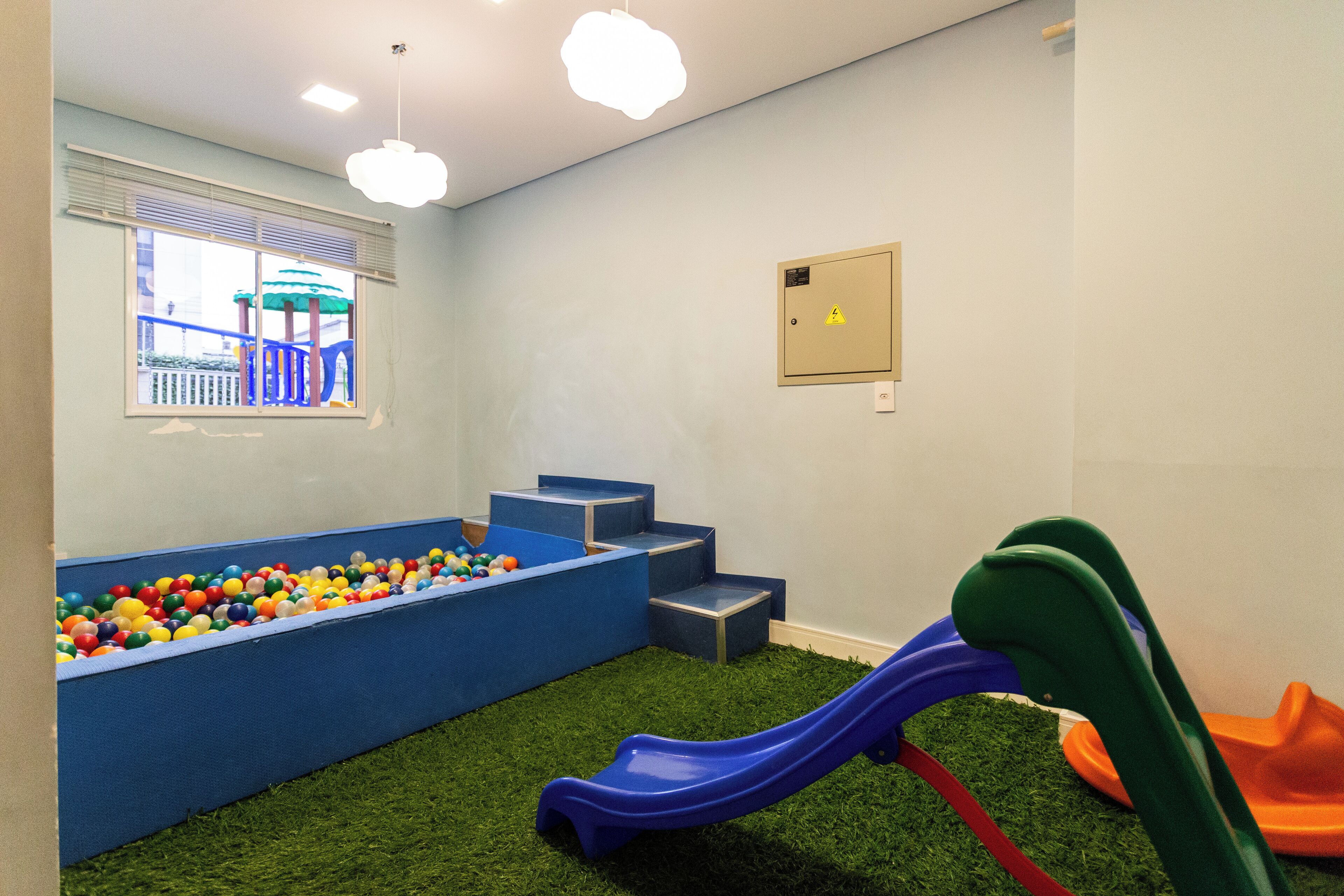A play area with a ball pit, a blue slide, and steps leading to a window overlooking a playground.
