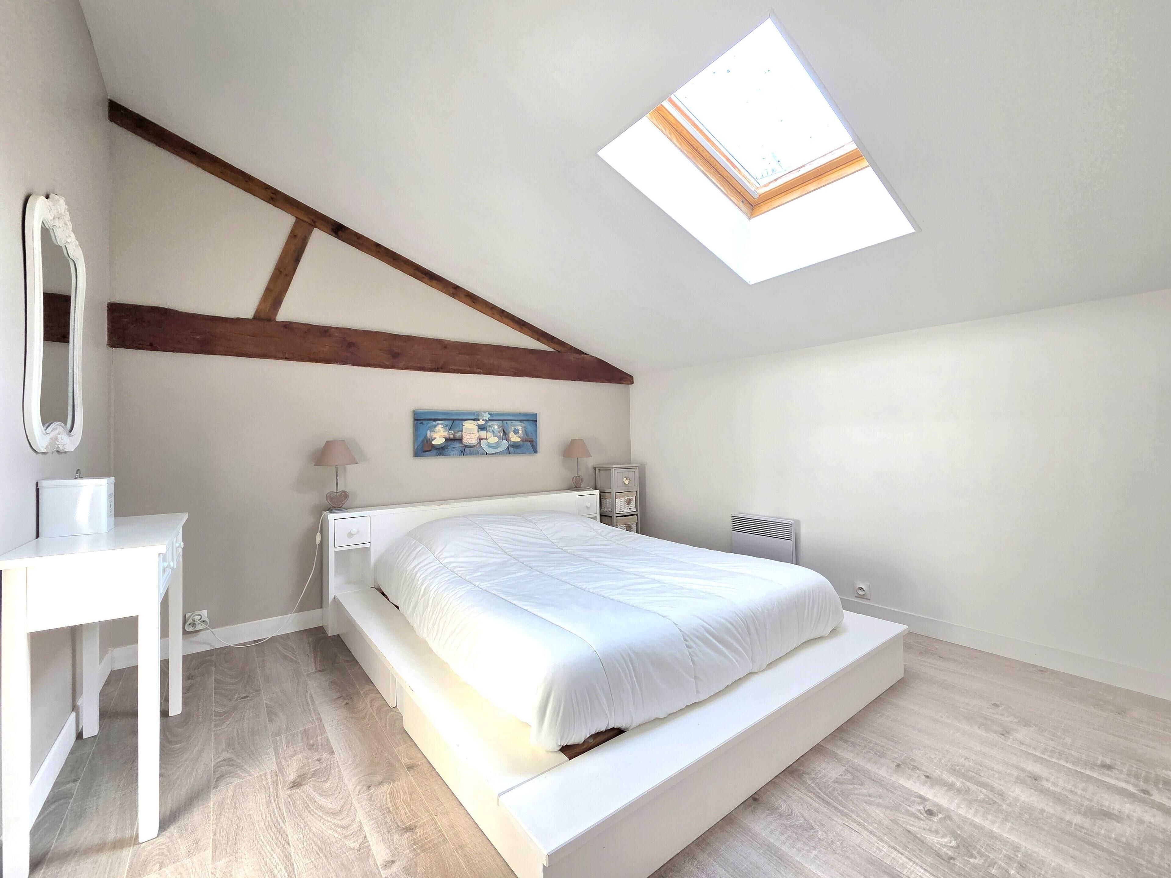 Une chambre avec un plafond en pente, un puits de lumière, un lit, une table de chevet, une commode, un miroir et une œuvre d’art accrochée au mur.