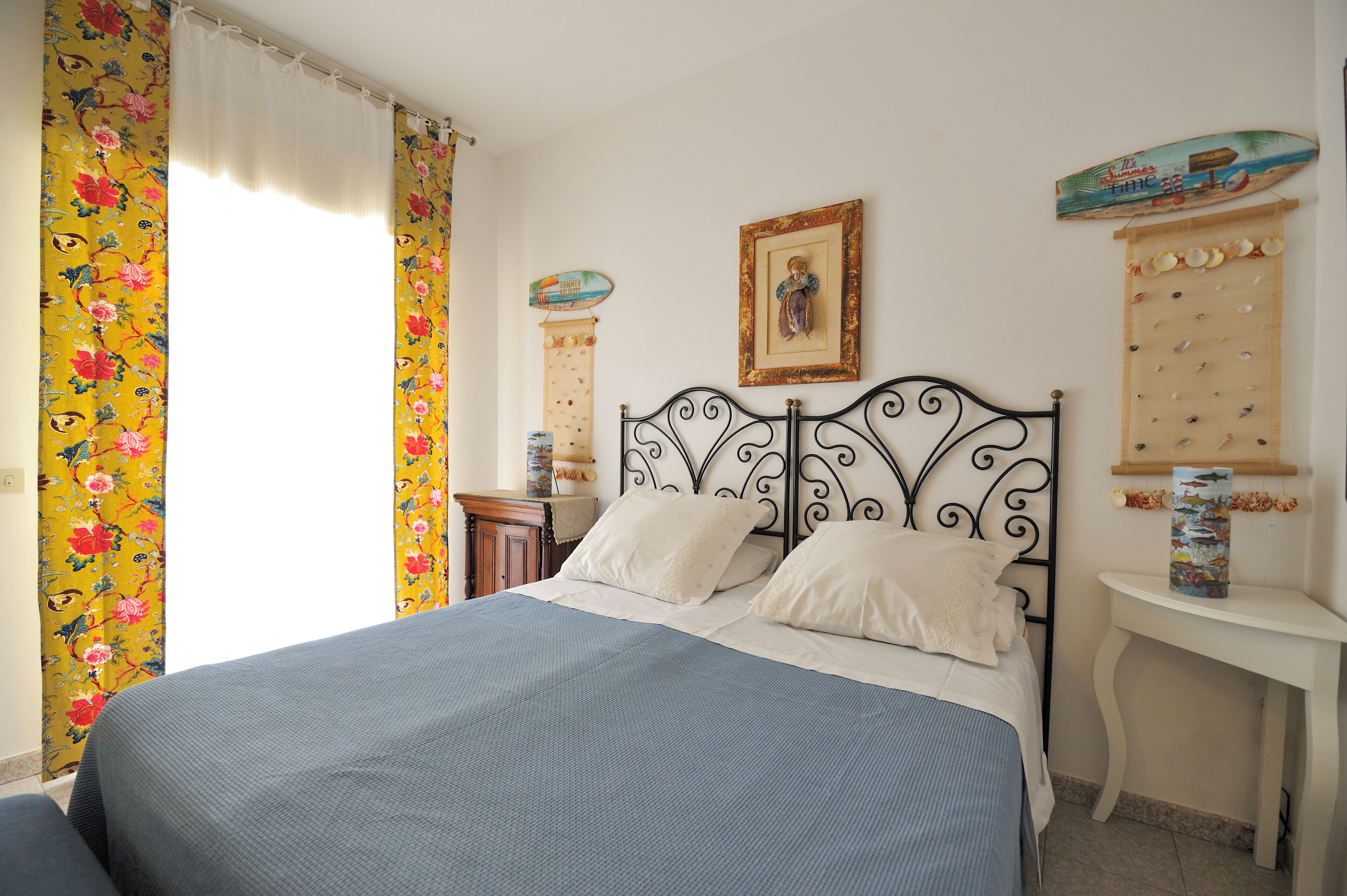 Una cama bien hecha con colcha azul, cabecero de metal negro y almohadas blancas. Hay una mesita de noche pequeña con un objeto decorativo. La habitación tiene cortinas con motivos florales y un cuadro en la pared.