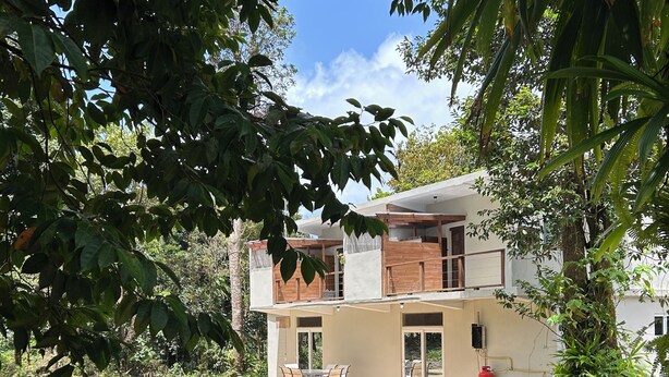 A two-story house with a balcony, surrounded by lush greenery and a clear sky.