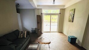 Uma sala de estar espaçosa com um sofá de cor escura, uma mesa de centro redonda e uma porta de vidro deslizante para uma área externa.