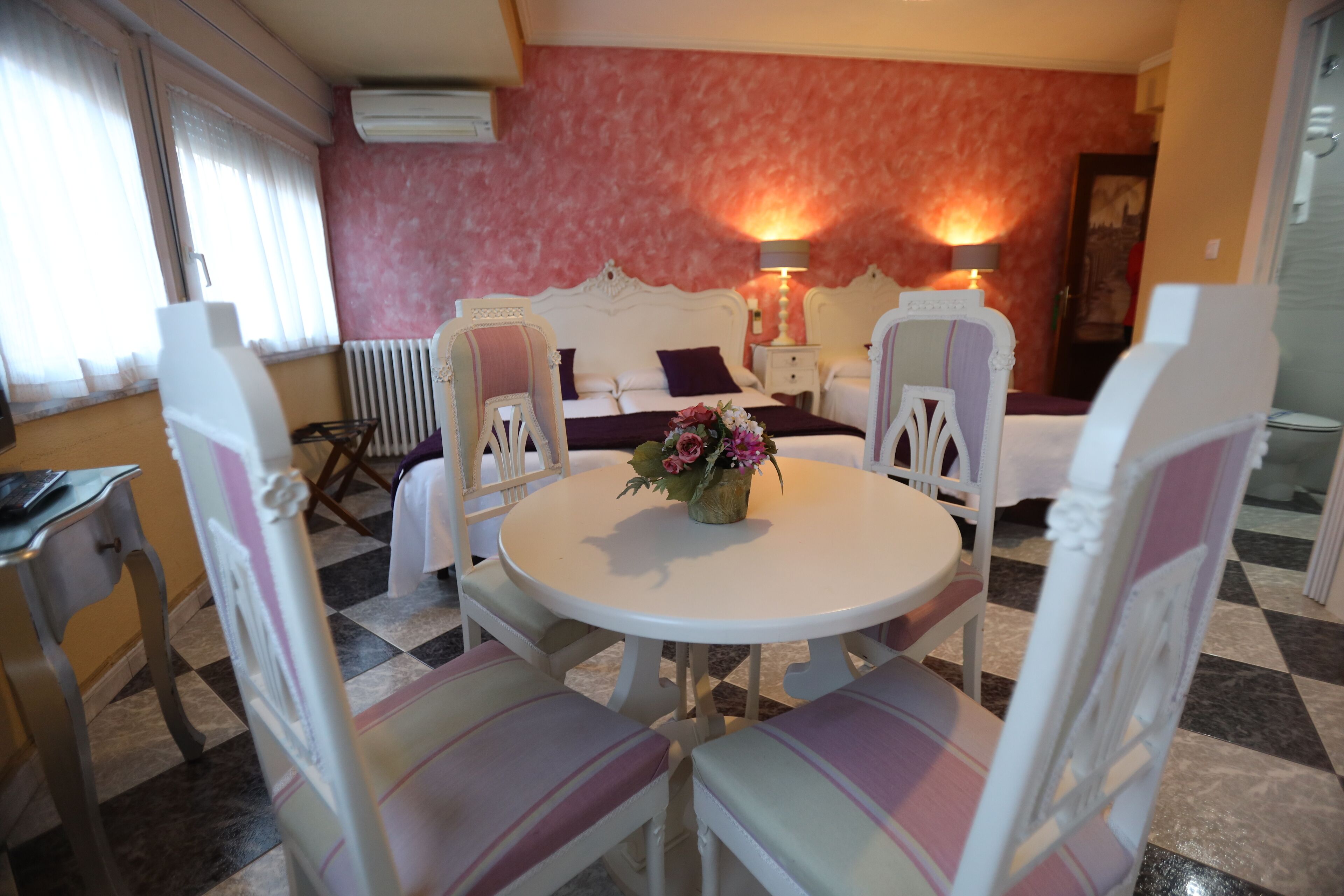 A room with a checkered floor, a round table with chairs, a bed with a headboard, and two bedside lamps.