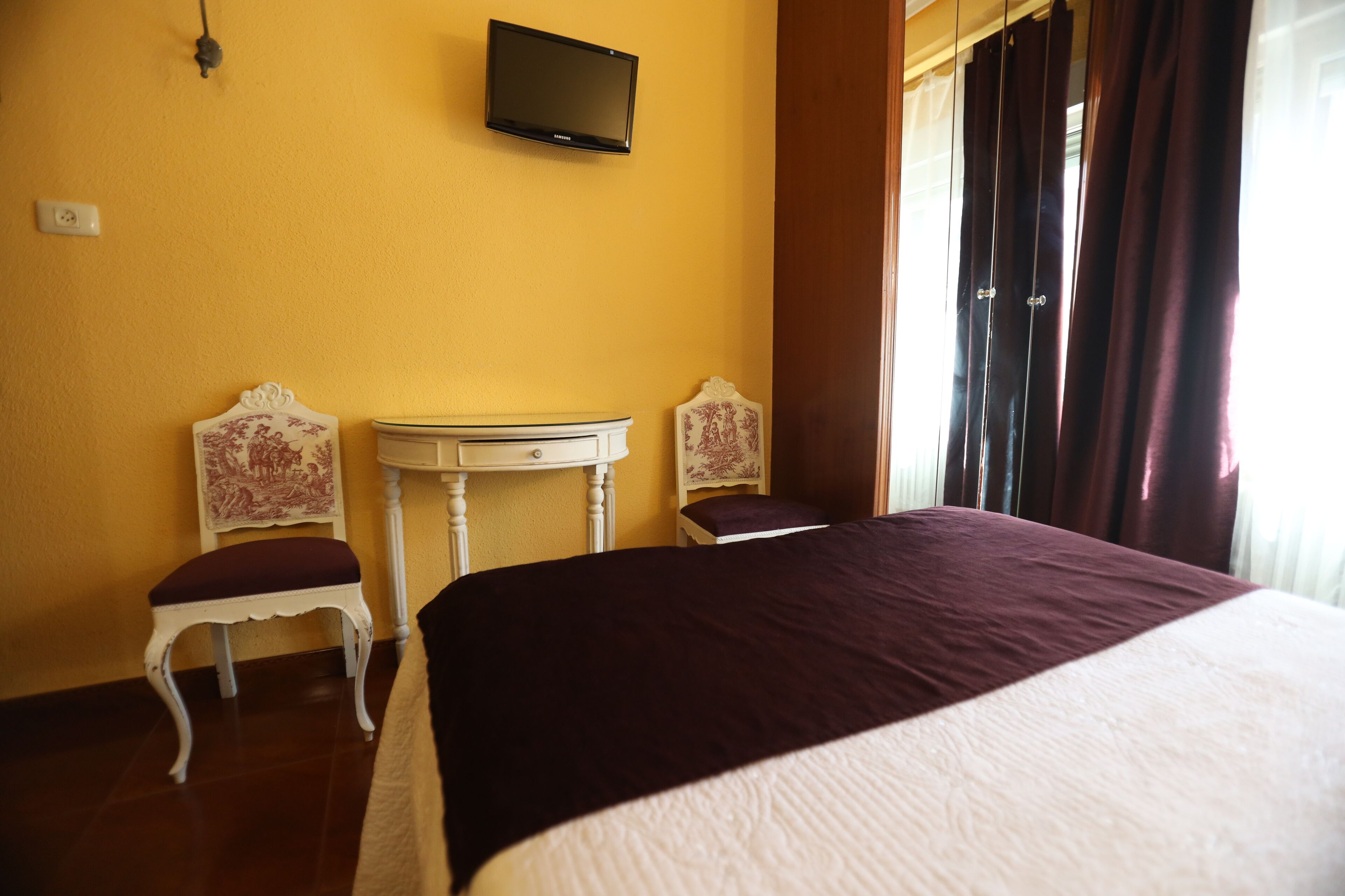 A hotel room with a bed, two chairs, a small table, a television on the wall, and a window with curtains.