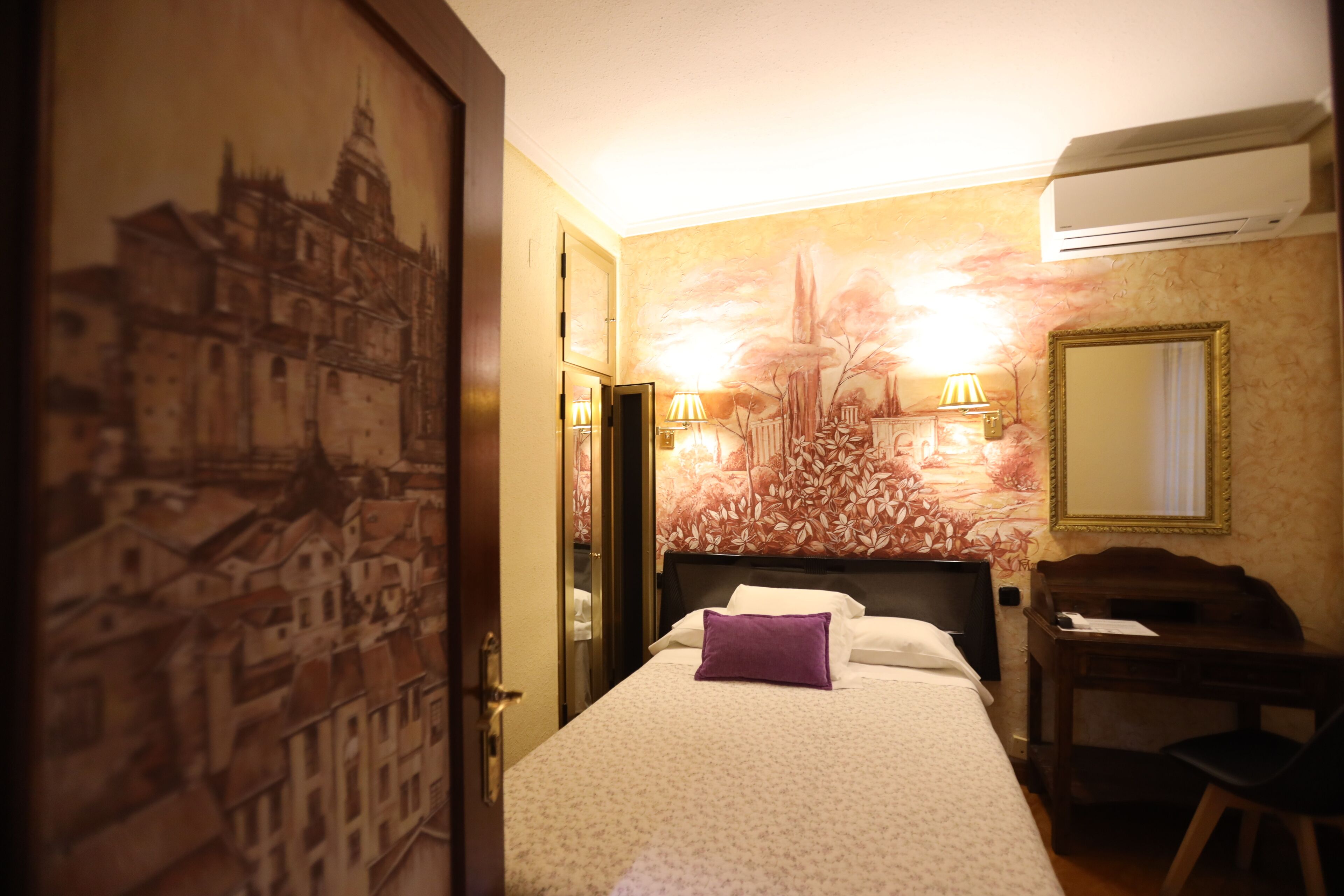 Habitación de hotel con cama, escritorio, silla, aire acondicionado montado en la pared y puerta con un mural de la ciudad.