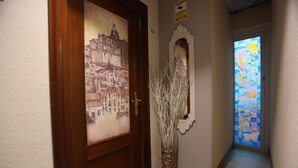 A hallway with a door featuring a cityscape painting, a vase with branches, and a stained glass window. - Hostal Goya Suites (Salamanca)