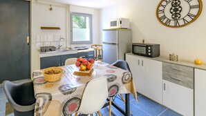 A compact kitchen with a dining table, chairs, a microwave, a refrigerator, and a large wall clock.