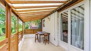 A covered patio area with a wooden table and two stools, a sliding glass door, and a view of the garden.