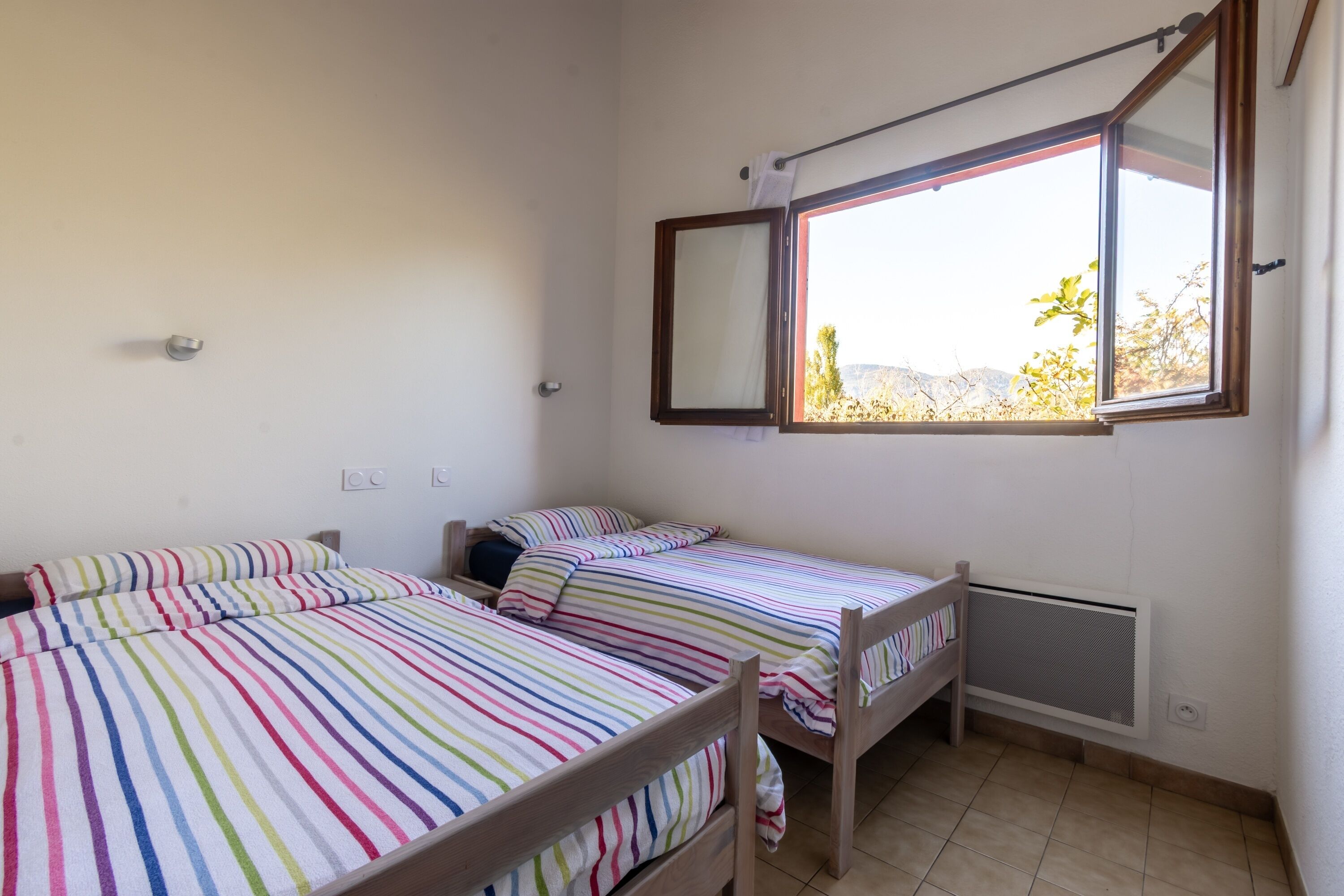 A room with two wooden beds, a window with a view of greenery, and a radiator on the wall.